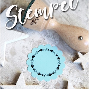 Könnte beinhalten: Ein Holzstempel mit dem Wort "Stempel" in weißer Schrift. Der Stempel hat ein grünes Stempelkissen und befindet sich neben einem hellblauen, gewellten Kreis mit einem schwarzen Herz- und Punktmuster. Weiße Holzsterne sind verstreut.