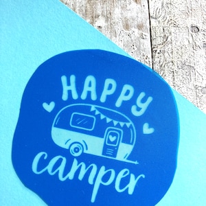 Könnte beinhalten: Blauer Kreis mit weißem Text, der "Happy Camper" sagt, und einer weißen Illustration eines Wohnwagens mit Flaggen und einem Herzen.