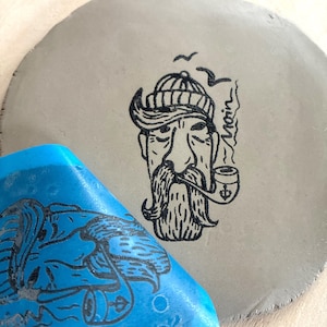 Könnte beinhalten: Ein grauer, runder Keramikteller mit einer schwarzen Illustration eines Seemanns, der eine Pfeife raucht. Der Seemann hat einen Schnurrbart und eine Strickmütze. Ein blaues, rechteckiges Objekt mit einem Elefantenmuster befindet sich im Vordergrund.