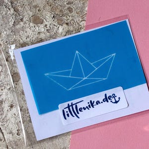 Könnte beinhalten: Eine blaue Karte mit einer weißen Papierboot-Illustration. Die Karte hat ein weißes Etikett mit dem Text "fittenika.de" und einem Ankersymbol. Die Karte befindet sich in einer transparenten Plastikhülle.