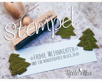 May include: A wooden stamp with the text "little nika" and the word "Stempel" written in cursive above it. The stamp is on a light brown surface with three green felt Christmas trees.