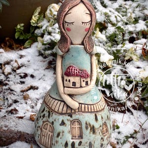 Könnte beinhalten: Keramikfigur eines Mädchens in einem blauen Kleid, das ein kleines Haus hält. Die Figur hat geschlossene Augen, rosige Wangen und braunes Haar. Das Kleid hat einen gemusterten Rock mit Tür- und Fensterdetails. Das Haus hat ein rotes Dach.