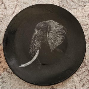 May include: A black ceramic plate featuring a detailed, grayscale illustration of an elephant's head and tusk. The elephant is depicted in profile, with textured details on the ear and trunk. The plate has a glossy finish.