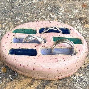 Pode incluir: Um porta-anéis de cerâmica rosa salpicada com quatro compartimentos em tons de azul e verde. O suporte contém quatro anéis de prata de vários designs. O porta-anéis tem formato oval e fica sobre uma superfície texturizada.