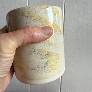 May include: A handmade ceramic mug with a cream-colored base, adorned with horizontal bands of yellow and speckled black detailing. The mug has a rounded shape and is held by a hand.