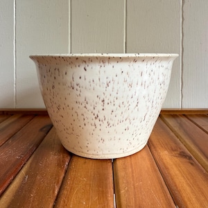 Peut inclure: Un bol en céramique moucheté avec un bord évasé. Le bol est blanc cassé avec de petites mouchetures brun foncé. Il est posé sur une surface en bois. L'arrière-plan est un mur de couleur claire.