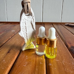 May include: A cream-coloured ceramic fish air freshener with a tan leather cord hangs above two small glass bottles with gold-coloured caps. One bottle contains a clear liquid, the other a yellow liquid. The items rest on a wooden surface.