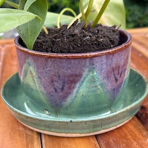 Puede incluir: Una maceta de cerámica hecha a mano con un platillo a juego. La maceta está esmaltada en tonos morados y verdes, con un diseño ondulado. Contiene tierra y una planta verde. El platillo es de color verde liso.