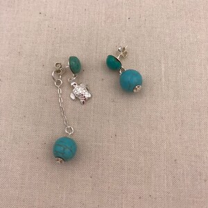 Puede incluir: Un par de pendientes de turquesa colgantes con un dije de tortuga de plata en uno de los pendientes. Los pendientes tienen una cuenta redonda de turquesa en la parte inferior.