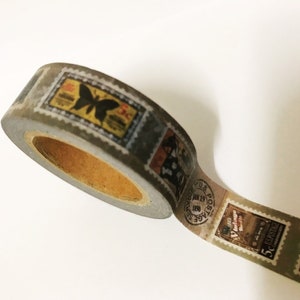 May include: A roll of washi tape featuring a vintage postage stamp design. The tape has a gray background with various stamp illustrations in shades of yellow, black, and brown. The tape is partially unrolled, revealing the inner cardboard core.
