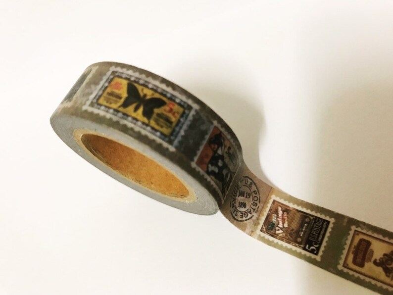 May include: A roll of washi tape featuring a vintage postage stamp design. The tape has a gray background with various stamp illustrations in shades of yellow, black, and brown. The tape is partially unrolled, revealing the inner cardboard core.