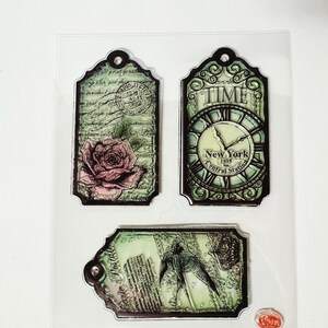 May include: Three vintage-style decorative tags in a green and black colour scheme. The first tag features a pink rose and a postage stamp. The second tag features a clock face with the text "TIME" and "New York 1895 Central Station". The third tag features a bird in flight and the text "Postage".