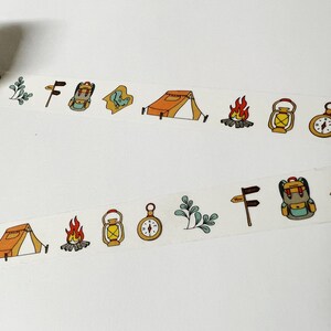 May include: A roll of washi tape with a repeating pattern of camping and outdoor adventure themed illustrations. The illustrations include a tent, a compass, a lantern, a backpack, a campfire, and a signpost.