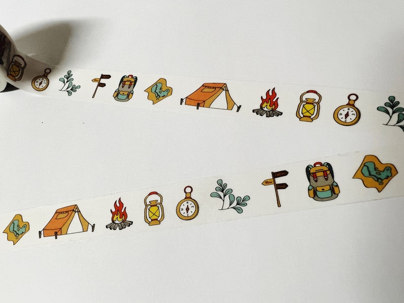 May include: A roll of washi tape with a repeating pattern of camping and outdoor adventure themed illustrations. The illustrations include a tent, a compass, a lantern, a backpack, a campfire, and a signpost.