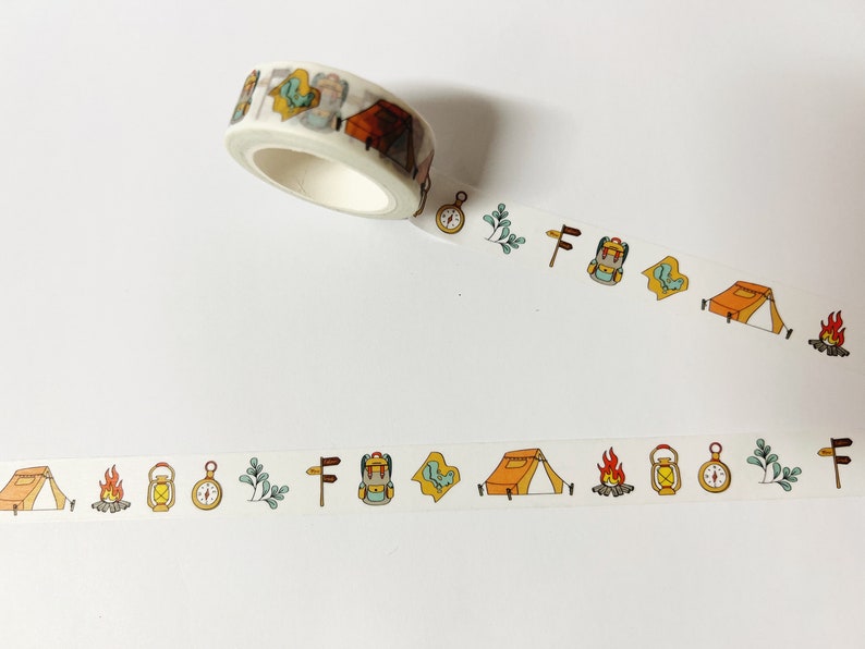 May include: A roll of white washi tape with a repeating pattern of camping-themed illustrations. The illustrations include a tent, a compass, a lantern, a backpack, a campfire, and a tree.