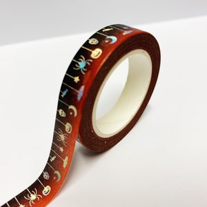 Op de afbeelding: Een rol decoratieve washi tape met een Halloween thema. De tape heeft een donkerbruine achtergrond met een herhalend patroon van spinnen, vleermuizen, manen en schedels in iriserende kleuren. De tape is ongeveer 1,5 cm breed.