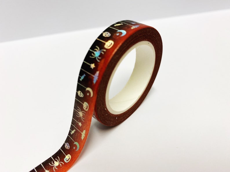 Op de afbeelding: Een rol decoratieve washi tape met een Halloween thema. De tape heeft een donkerbruine achtergrond met een herhalend patroon van spinnen, vleermuizen, manen en schedels in iriserende kleuren. De tape is ongeveer 1,5 cm breed.