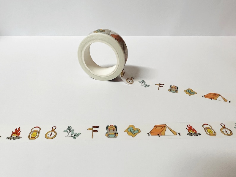 May include: A roll of white washi tape with a camping theme. The tape features illustrations of a tent, a compass, a lantern, a backpack, a campfire, and a signpost.