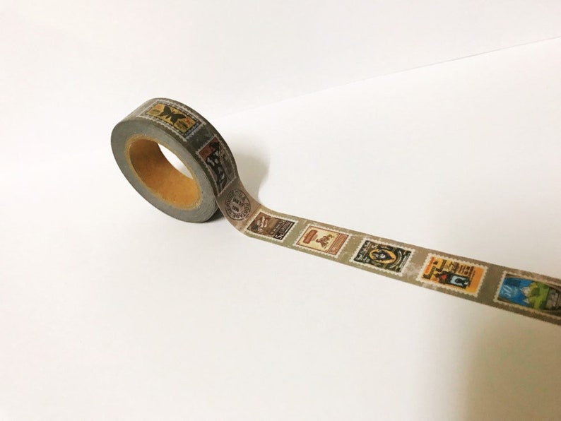 May include: A roll of decorative washi tape featuring a repeating pattern of vintage postage stamps in various colors. The tape is gray with a brown core and is unrolled on a white surface.
