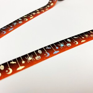Op de afbeelding: Twee stroken oranje en bruine decoratieve tape met Halloween-thema. De tape is versierd met zilveren spinnen, pompoenen, manen en sterren. De tape staat op een witte achtergrond.