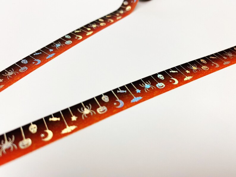 Op de afbeelding: Twee stroken oranje en bruine decoratieve tape met Halloween-thema. De tape is versierd met zilveren spinnen, pompoenen, manen en sterren. De tape staat op een witte achtergrond.