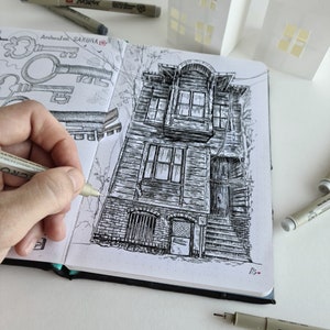 May include: Detailed pen and ink drawing of a weathered building in a sketchbook. The drawing is in black and white, with intricate details of the building's facade. A hand is holding a pen, adding to the artwork. The sketchbook is open, revealing other sketches.