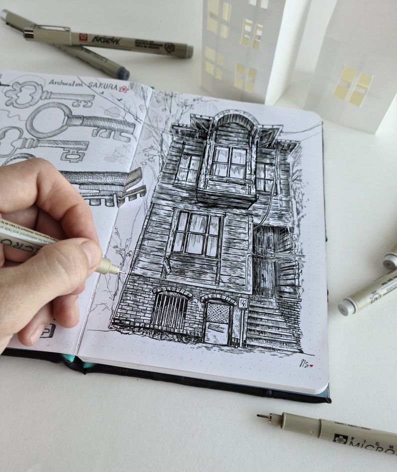 May include: Detailed pen and ink drawing of a weathered building in a sketchbook. The drawing is in black and white, with intricate details of the building's facade. A hand is holding a pen, adding to the artwork. The sketchbook is open, revealing other sketches.