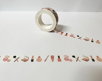 May include: A roll of decorative washi tape featuring a repeating pattern of makeup items in shades of pink, brown, and beige. The tape includes images of lipstick, eyeshadow, and brushes.