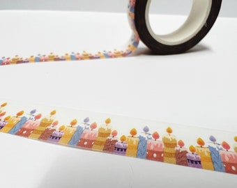 May include: Decorative washi tape featuring a repeating pattern of colorful birthday cakes with lit candles. The tape roll is visible, with the patterned tape extending across a white surface. The cakes have various colors, including yellow, pink, and purple.
