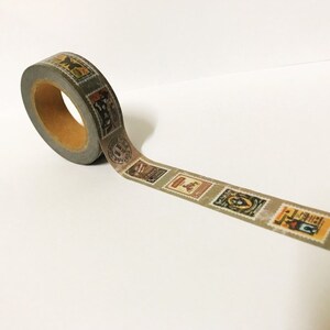 May include: A roll of decorative washi tape featuring a repeating pattern of vintage postage stamp designs in shades of brown, tan, and green. The tape is gray with a brown core.