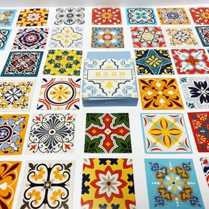 May include: A display of square tiles featuring detailed floral and geometric designs in shades of blue, yellow, red, and green. A small box with Chinese text is placed centrally on the tiles.