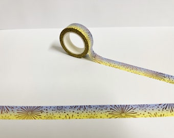 May include: A roll of decorative washi tape with a fireworks design. The tape features a gradient of yellow and blue with gold-colored fireworks and starburst patterns. The tape is unrolled on a white surface.