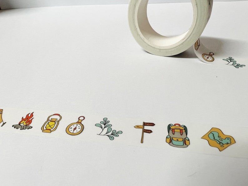 May include: A roll of washi tape with a camping theme. The tape features illustrations of a tent, campfire, lantern, compass, leaves, a signpost, a backpack, and a map.