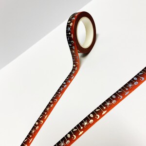 Op de afbeelding: Een rol Halloween-thema washi tape. De tape heeft een bruin tot oranje verloop met een herhalend patroon van spinnen, manen, vleermuizen en pompoenen in zilver en blauw. De tape is afgerold.
