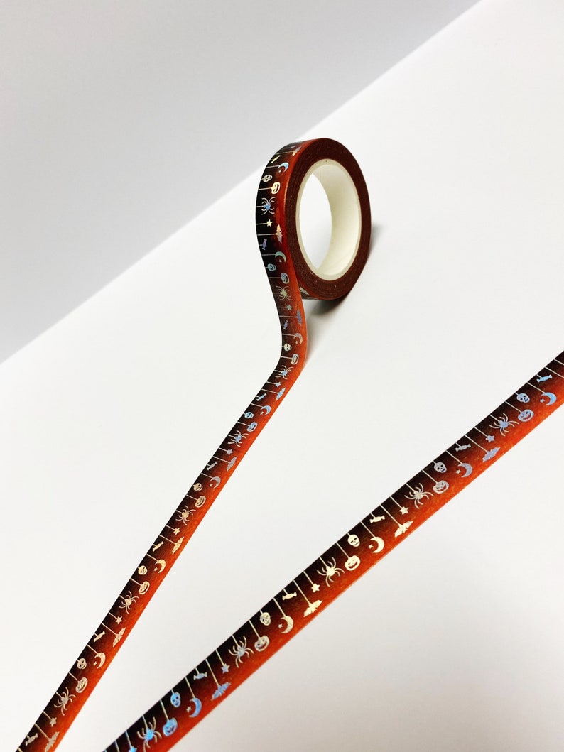 Op de afbeelding: Een rol Halloween-thema washi tape. De tape heeft een bruin tot oranje verloop met een herhalend patroon van spinnen, manen, vleermuizen en pompoenen in zilver en blauw. De tape is afgerold.