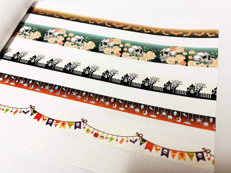 Op de afbeelding: Halloween-thema washi tape rollen. Ontwerpen omvatten vleermuizen, schedels met rozen, spookhuizen, spinnen, manen en feestelijke banners in oranje, zwart en groen. Perfect voor knutselen en scrapbooking.