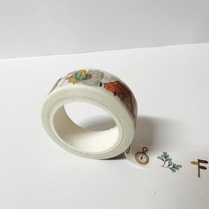 May include: A roll of white washi tape with a camping theme. The tape features illustrations of a tent, a compass, a backpack, and a tree. The tape is partially unrolled, showing the design.