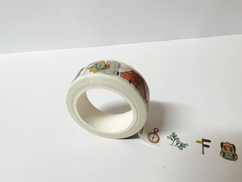 May include: A roll of white washi tape with a camping theme. The tape features illustrations of a tent, a compass, a backpack, and a tree. The tape is partially unrolled, showing the design.