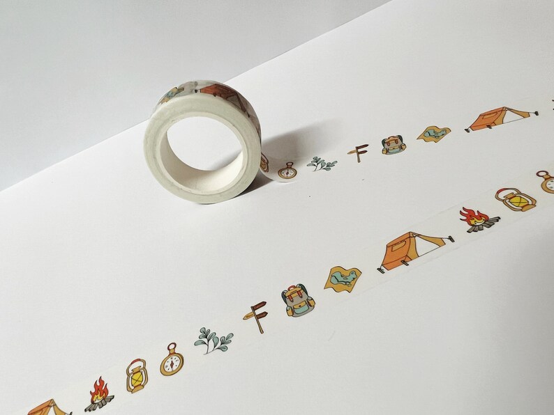 May include: A roll of white washi tape with a camping theme. The tape features illustrations of tents, campfires, compasses, backpacks, and trees.