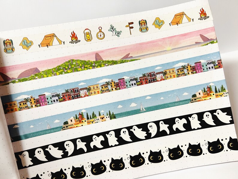 May include: A collection of six colorful washi tapes featuring various designs, including a camping scene, a cityscape, a seascape, ghosts, and cats. The tapes are all on a white background.