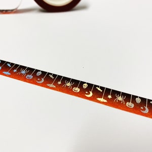 Op de afbeelding: Halloween-washi-tape met een herhalend patroon van spinnen, pompoenen, manen, vleermuizen en sterren. De tape heeft een verlopende oranje en zwarte achtergrond met metallic zilveren accenten. Een rol van dezelfde tape is zichtbaar.