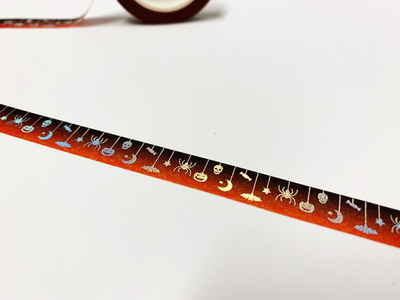 Op de afbeelding: Halloween-washi-tape met een herhalend patroon van spinnen, pompoenen, manen, vleermuizen en sterren. De tape heeft een verlopende oranje en zwarte achtergrond met metallic zilveren accenten. Een rol van dezelfde tape is zichtbaar.