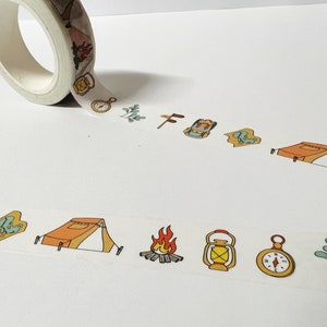 May include: A roll of washi tape with a camping theme. The tape features illustrations of a tent, a compass, a lantern, a backpack, a campfire, and a leaf.