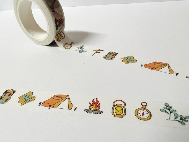 May include: A roll of washi tape with a camping theme. The tape features illustrations of a tent, a compass, a lantern, a backpack, a campfire, and a leaf.