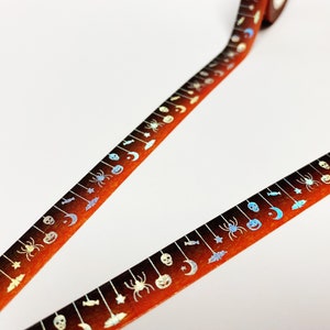 Op de afbeelding: Halloween-thema washi tape met een herhalend patroon van spinnen, vleermuizen, manen en pompoenen. De tape heeft een oranje en bruine gradi&euml;nt achtergrond met zilveren accenten. Perfect voor knutselen en scrapbooking.