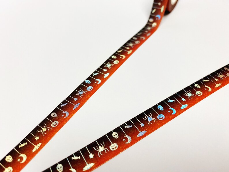 Op de afbeelding: Halloween-thema washi tape met een herhalend patroon van spinnen, vleermuizen, manen en pompoenen. De tape heeft een oranje en bruine gradi&euml;nt achtergrond met zilveren accenten. Perfect voor knutselen en scrapbooking.
