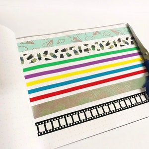 May include: A white notebook with a dotted pattern and a variety of colourful washi tapes, including a green tape with a cactus pattern, a purple tape, a yellow tape, a blue tape, a red tape, a silver tape, and a black and white film strip tape. A pair of blue scissors is also in the image.