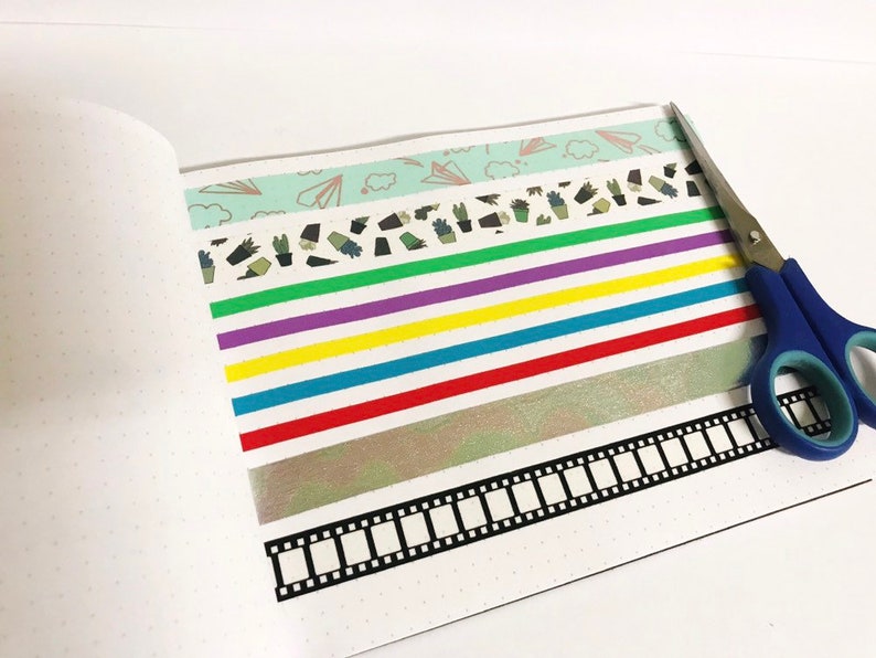 May include: A white notebook with a dotted pattern and a variety of colourful washi tapes, including a green tape with a cactus pattern, a purple tape, a yellow tape, a blue tape, a red tape, a silver tape, and a black and white film strip tape. A pair of blue scissors is also in the image.