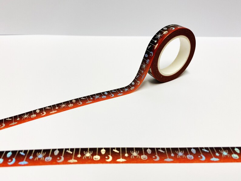 Op de afbeelding: Een rol Halloween washi tape met een oranje en bruine gradi&euml;nt achtergrond. De tape heeft zilveren illustraties van spinnen, pompoenen, manen en andere griezelige elementen. De tape is afgerold en toont het herhalende patroon.