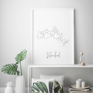 以下が含まれることがあります： イスタンブールの輪郭の黒い線画が描かれた、額入りのミニマリストアートプリント。 「Istanbul」の文字が筆記体で下に書かれています。 クッションやパイナップル型の花瓶など、装飾品が置かれた白い棚に飾られています。